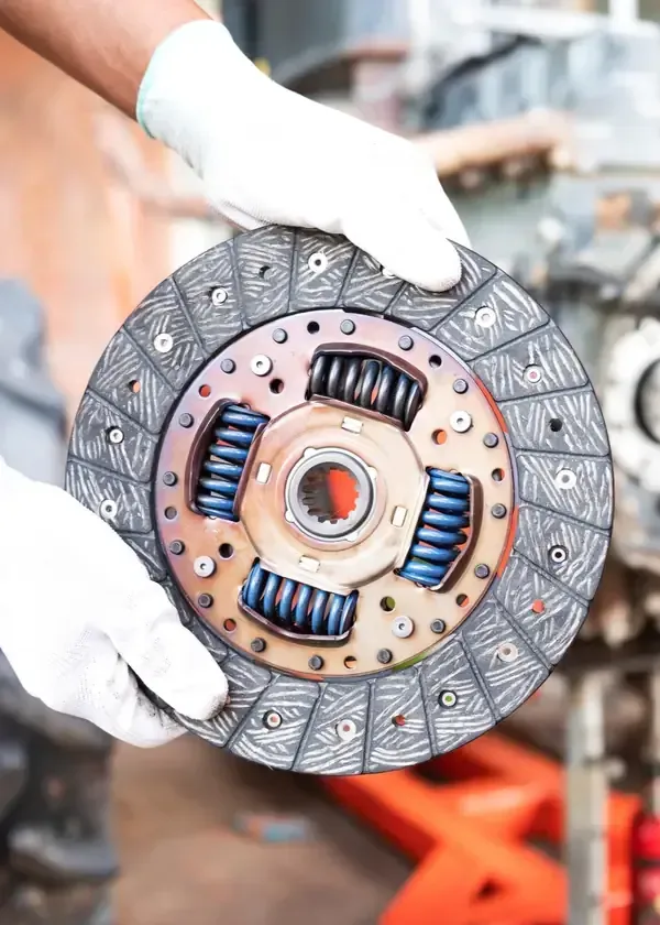 Hands in white gloves hold a circular automotive clutch disc with visible blue springs in a workshop.