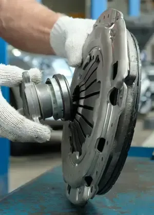 Hands in work gloves holding a metallic automotive clutch pressure plate and release bearing in a workshop.