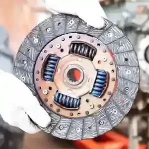 Hands in white gloves hold a circular automotive clutch disc with visible blue springs in a workshop.
