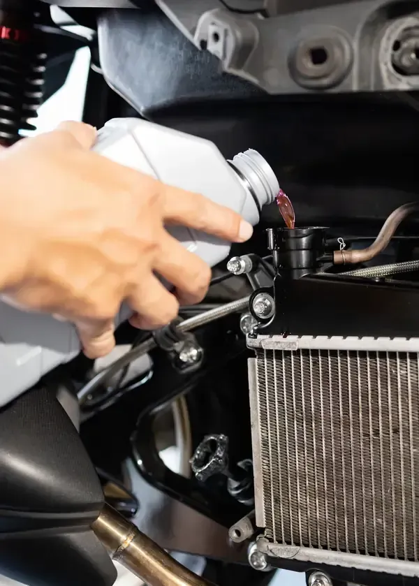 A person pours red liquid from a bottle into the radiator reservoir of a motorcycle engine.