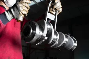 A worker in a red shirt and protective gloves holds a metal crankshaft using a specialized lifting tool.