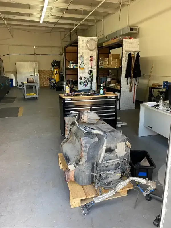 A large, dark vehicle engine sits on a wooden pallet in the middle of a brightly lit industrial workshop or garage.