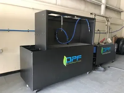 A black DPF cleaning machine for diesel particulate filters sits in a workshop with blue hoses and a logo on the front.