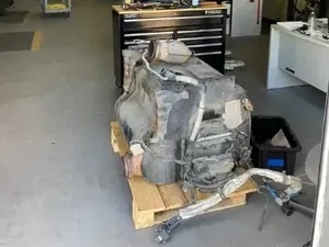 A large, dark vehicle engine sits on a wooden pallet in the middle of a brightly lit industrial workshop or garage.