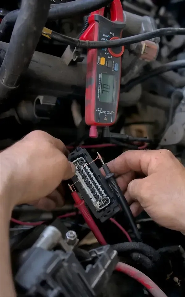 Hands test a vehicle’s wiring harness connector using a red multimeter clamped to a wire and a black probe on the pins.