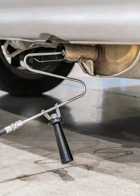 A metal emissions testing probe inserted into a car's exhaust pipe, connected to a hose with a black handle on the ground.
