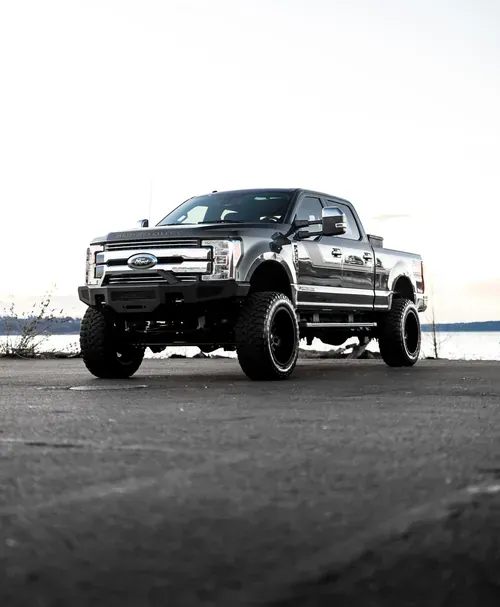 A dark-colored, lifted Ford Super Duty pickup truck parked on asphalt near a body of water at sunset.