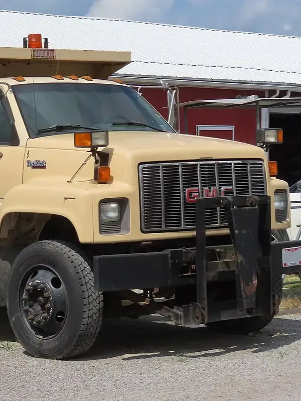 A tan GMC dump truck with a front-mounted snowplow attachment, parked on a gravel lot in front of a red building.
