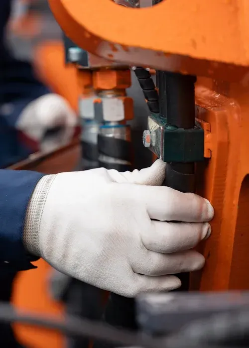 A hand in a white glove holds a black hose against orange industrial machinery.
