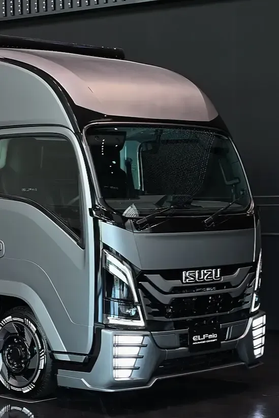 A silver Isuzu Elf light-duty truck viewed from the front-quarter angle against a dark background.