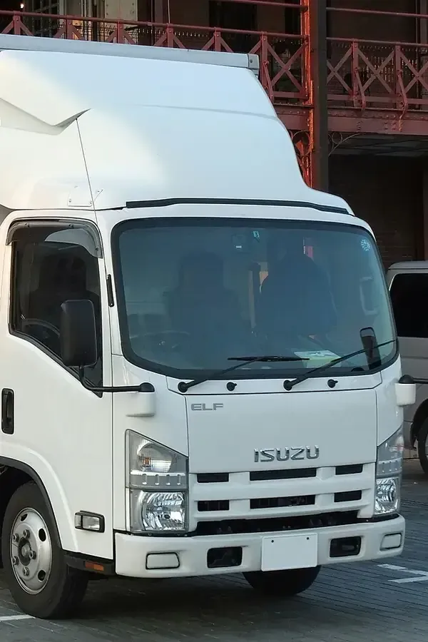 A white Isuzu Elf truck parked outdoors with a partially visible building structure in the background.