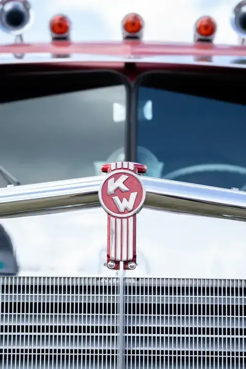 A close-up of a vintage Kenworth truck’s red and chrome hood emblem above a silver grille and rooftop amber lights.