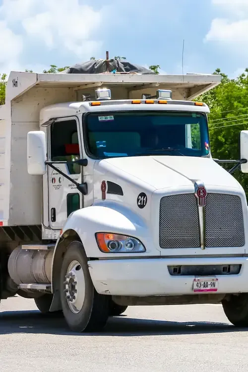 A white Kenworth dump truck driving on a paved road with green trees in the background.