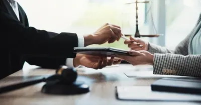 A lawyer in a suit and a client exchange a document across a desk, with a judge’s gavel and scales of justice visible.