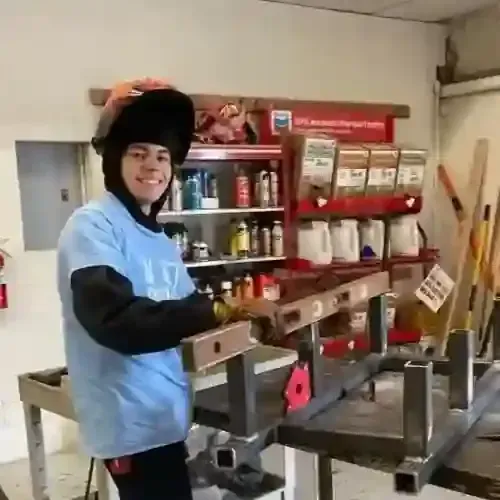 A person wearing a welding helmet and blue long-sleeve shirt works with metal components on a workbench in a workshop.