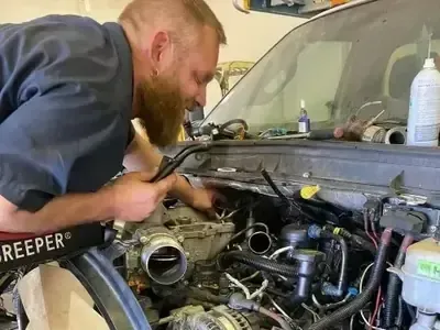 A mechanic with a beard works on the exposed engine of a vehicle in a repair shop.
