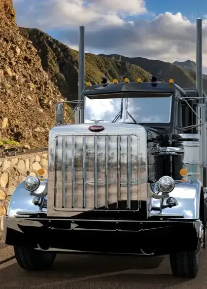 A classic black Peterbilt semi-truck with a large chrome grille and exhaust stacks parked near a rocky mountain roadside.