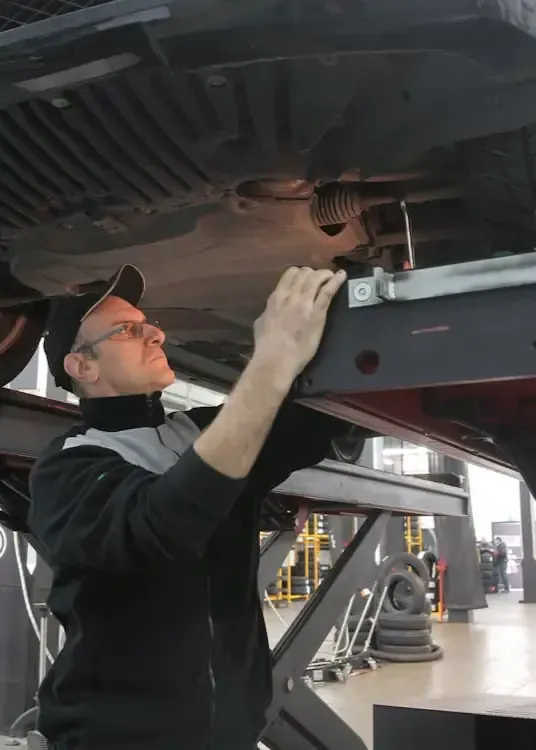 A technician in a black and grey jacket uses a car lift to inspect the underside of a vehicle in a repair shop.