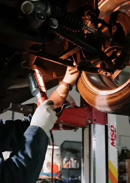 A mechanic uses a bright work light to inspect the undercarriage of a car raised on a hydraulic lift in a garage.