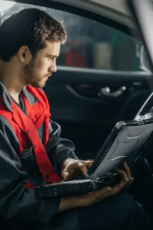 A mechanic in a red and grey uniform sits inside a vehicle, using a rugged laptop to diagnose car issues.