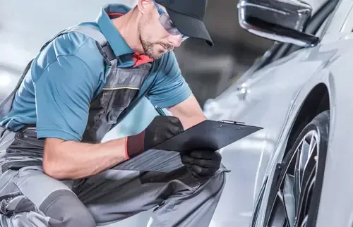 A mechanic in gray coveralls and safety glasses kneels by a white car, writing on a clipboard.