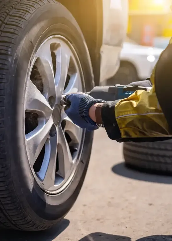 A worker wearing a safety jacket and blue gloves uses an impact wrench to tighten the lug nuts on a car wheel.