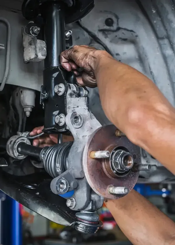 A mechanic's hands work on the front suspension and wheel hub assembly of a car in a repair shop.