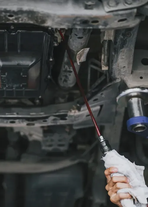 A hand holding a white cloth wipes a thin, red dipstick against the underside of a car engine.