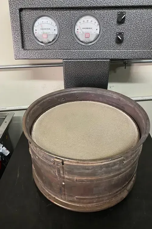A laboratory sieve shaker machine with a round metal sieve mounted on its platform under a control panel with two gauges.