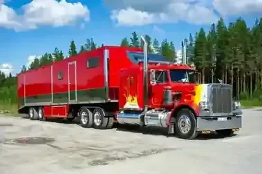 A bright red semi-truck with flame decals on the hood, hauling a long red trailer, parked on a dirt lot near a forest.