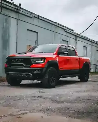 A bright red Ram TRX pickup truck parked on a gravel lot in front of a weathered, grey industrial building.