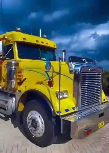 A bright yellow Freightliner semi-truck parked on a cobblestone surface under a dramatic, dark, cloudy sky.