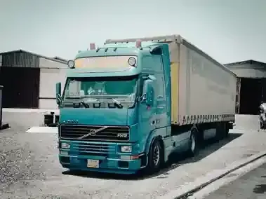 A teal Volvo semi-truck parked in front of industrial warehouse buildings on a sunny day.