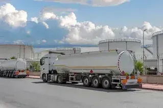Two white tanker trucks parked on a road next to large industrial fuel storage tanks under a cloudy sky.