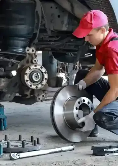 A mechanic wearing a red cap and shirt kneels while holding a brake disc in an auto repair shop.