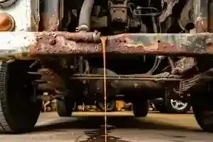 A rusty, vintage vehicle sits in a garage, leaking a stream of thick, orange liquid from a hose onto the concrete floor.