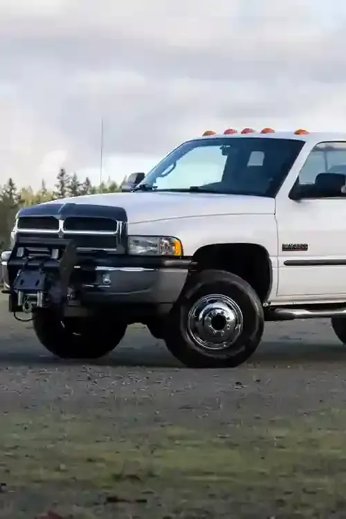 A white Dodge Ram dually pickup truck with a front-mounted winch, parked on a gravel surface under a cloudy sky.