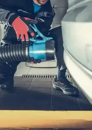 A technician in a workshop holds an exhaust extraction hose near the rear of a car.