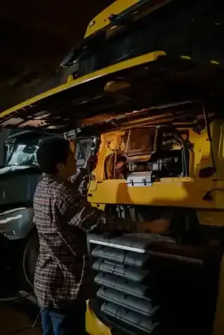 A person in a plaid shirt works on the open engine of a large yellow truck inside a dimly lit workshop.