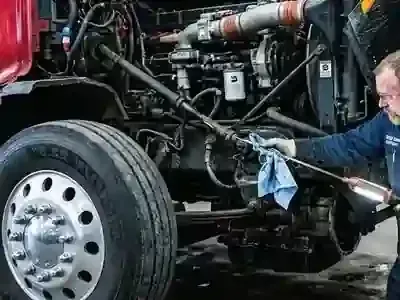 A mechanic wearing glasses wipes down the steering linkage of a red semi-truck engine with a blue shop rag.