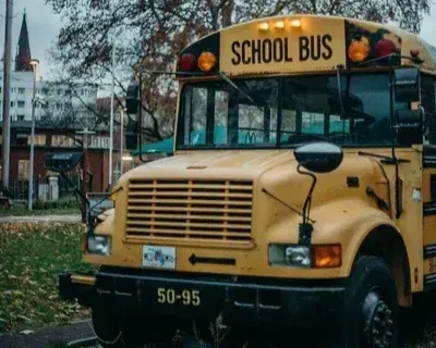 A yellow school bus is parked on a street with a tall building and trees in the background under a cloudy sky.