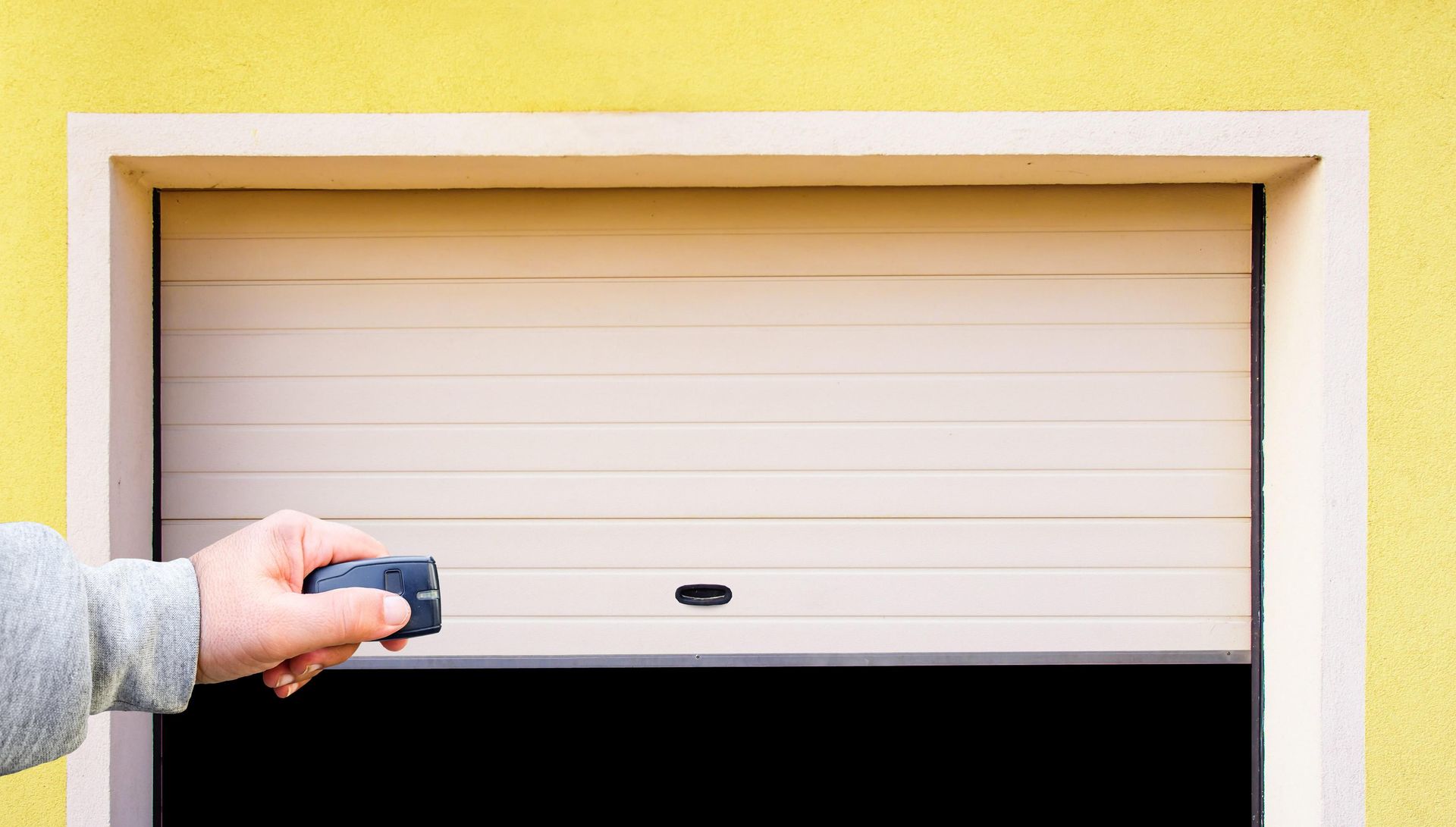 Hand using remote to open a tan, horizontal rolling shutter door set in a beige frame against a yellow wall.