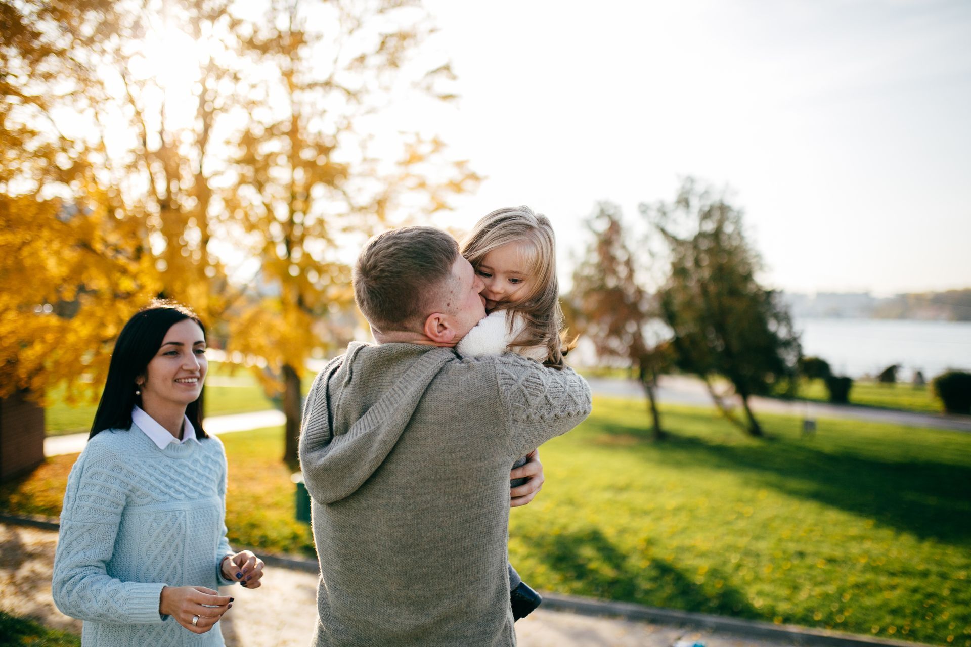 A man is holding a little girl in his arms in a park while a woman looks on.