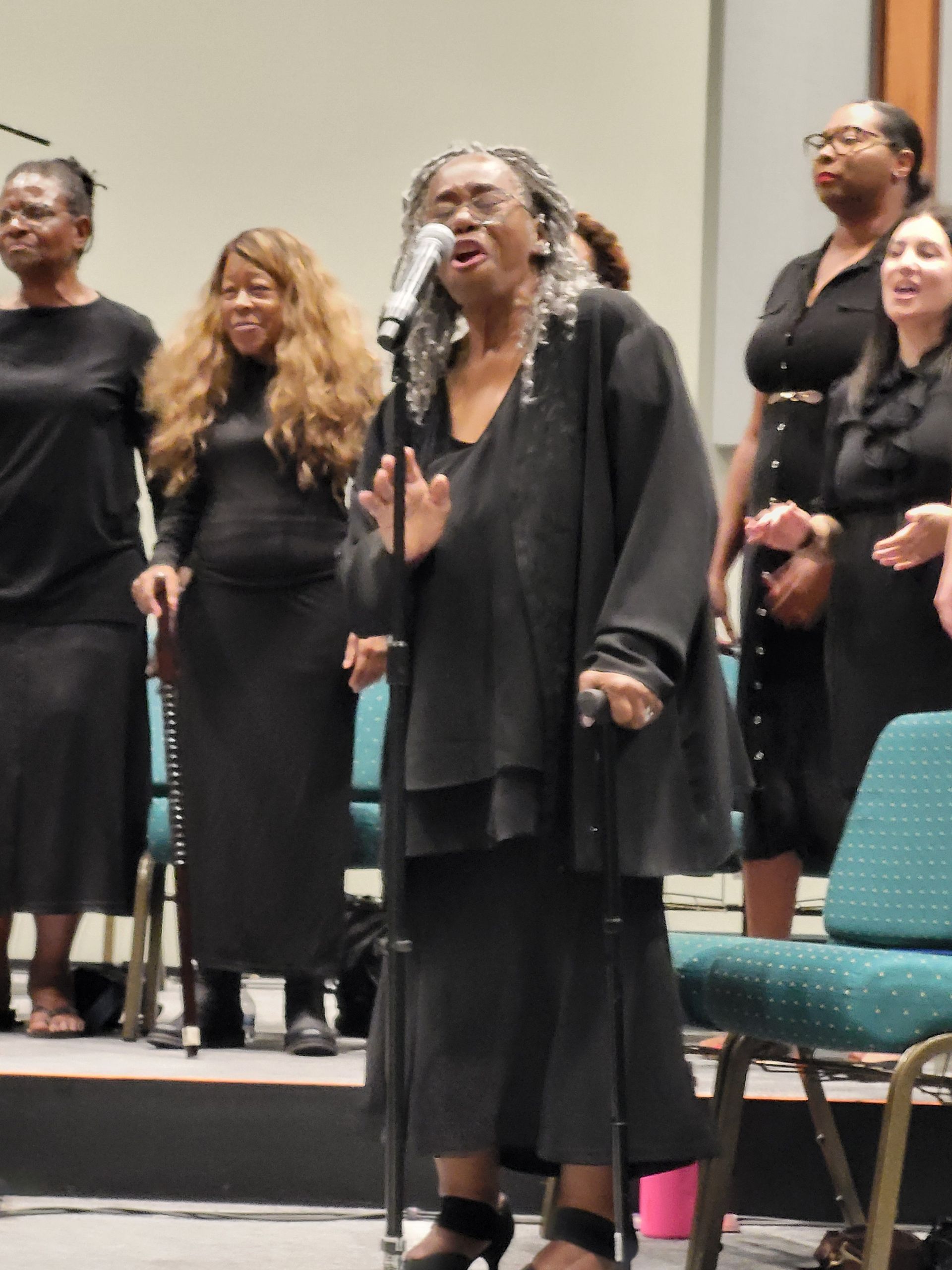 Choir singing on stage; lead singer in black, holding a microphone. Other choir members in black outfits.