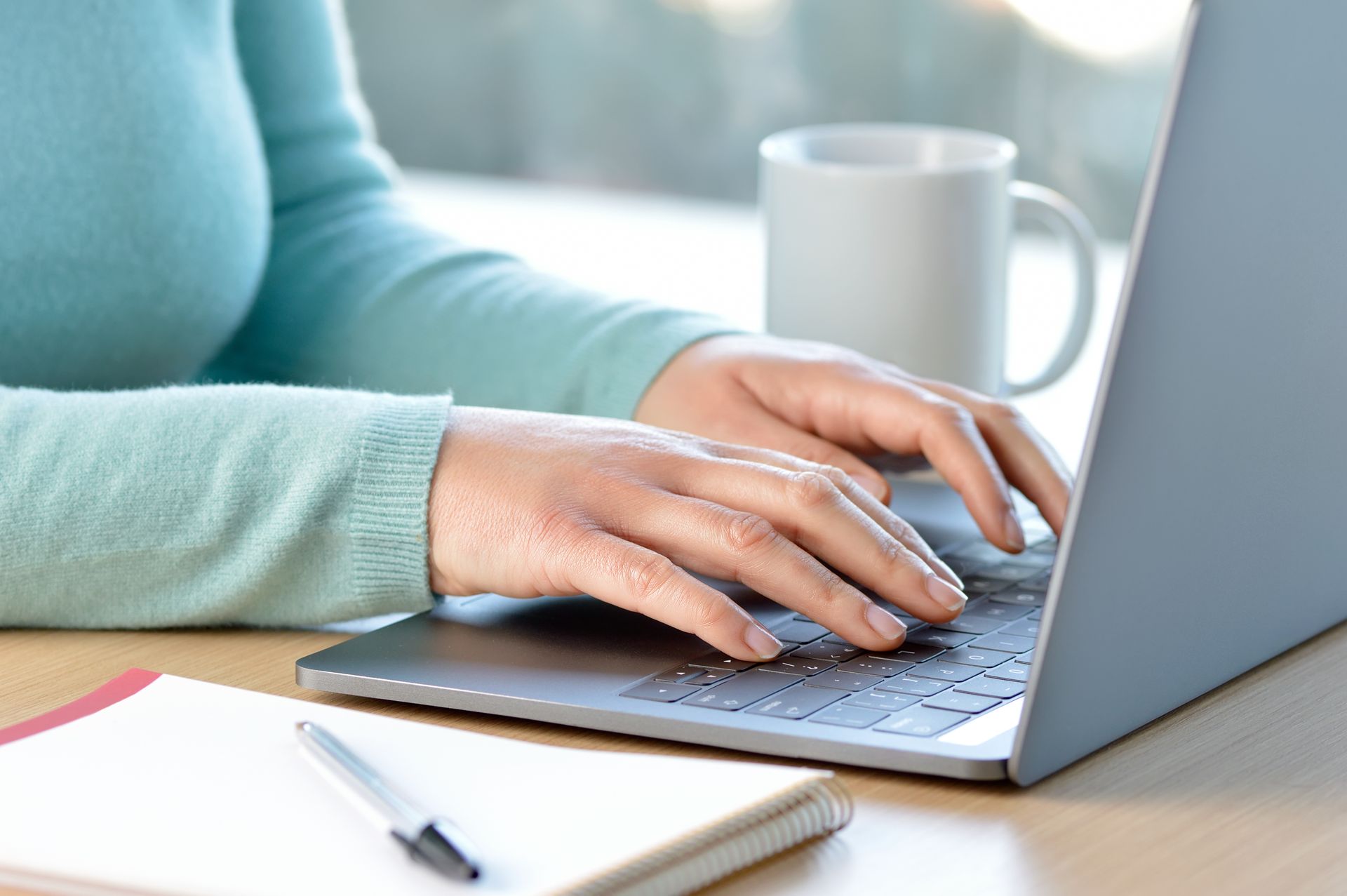 Person typing on a laptop with hands visible, notepad, and coffee mug on desk.