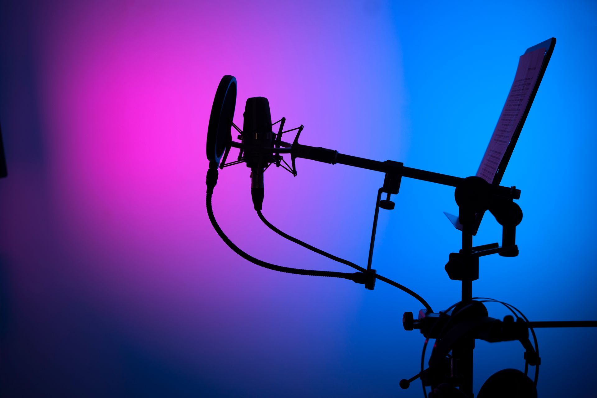 Silhouette of a microphone with pop filter, boom arm, and stand against a pink and blue lit background.