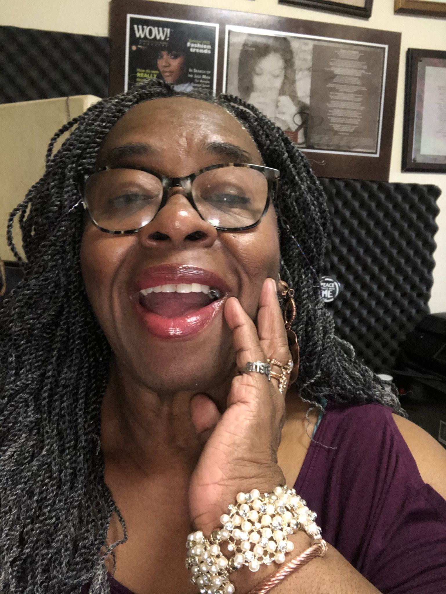 Woman with glasses smiles widely, hand on face, wearing braids and bracelets.