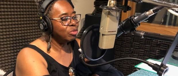 Person wearing headphones and glasses, speaking into a microphone in a recording studio.