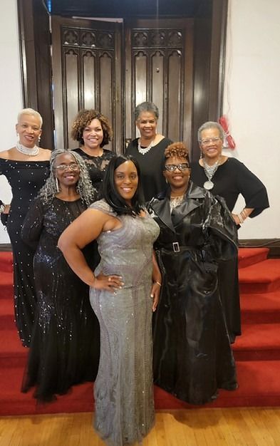 Group of women in formal gowns posing in front of wooden doors with a red carpet.