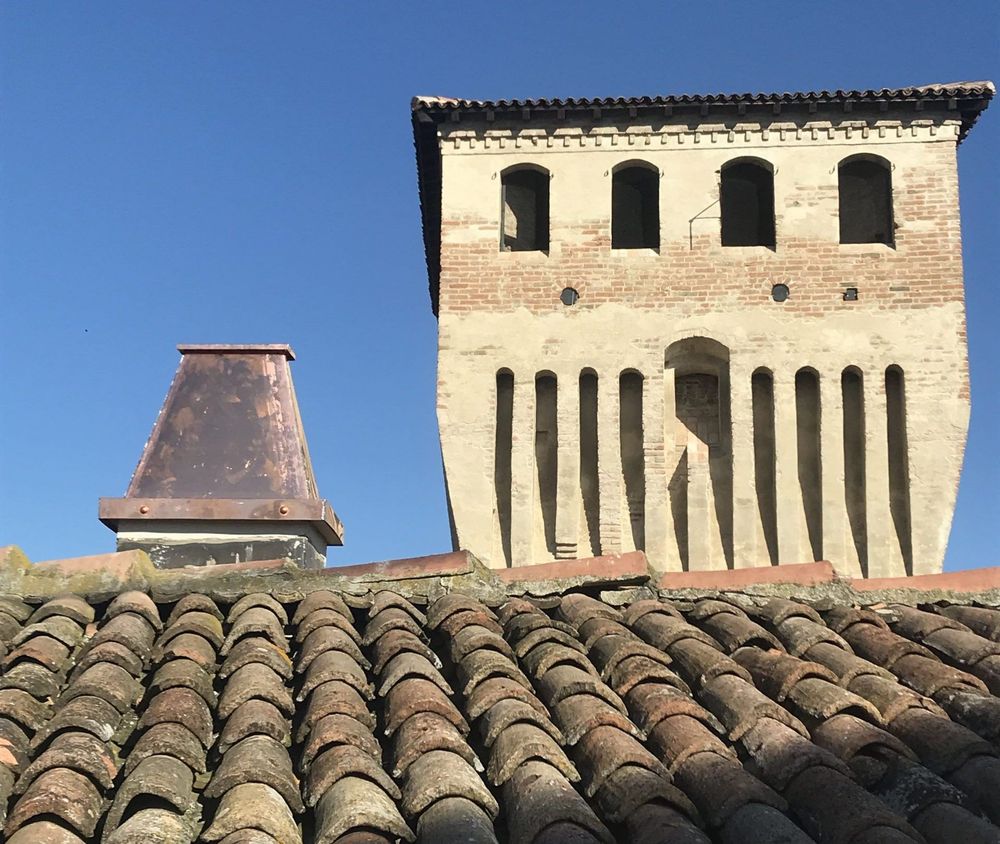Antica torre in pietra con finestre ad arco dietro le tegole del tetto in terracotta, sotto un cielo azzurro e limpido.